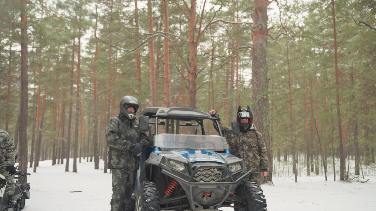 Due piloti accanto a un ATV blu in una pineta innevata. Conducenti con casco sono in piedi vicino a un veicolo utilitario (UTV) e a una motoslitta. I tronchi degli alberi sempreverdi e la neve fresca creano un'atmosfera selvaggia, con tracce di pneumatici visibili.