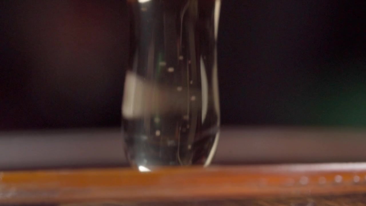 Vista macro di una goccia di miele che pende da un mestolo per miele sopra un barattolo di vetro, evidenziando la consistenza viscosa, la tensione superficiale, il colore ambrato e i dettagli riflettenti.
