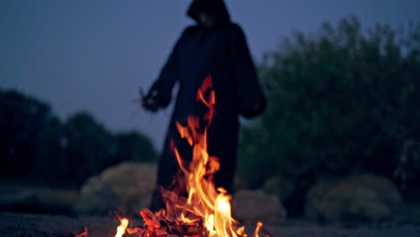 dark witch near fire. human in the coat with a hood on his head standing by the fire