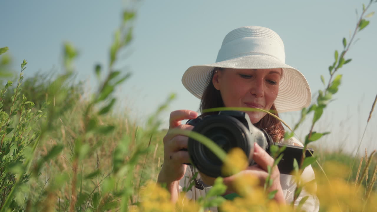 Excited woman wearing white hat squats in tall grass smiling while taking close-up photo of wildflowers in sunny green field surrounded by natural vegetation under clear blue sky