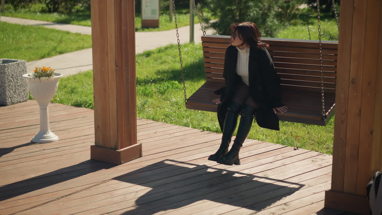 세련된 검은 코트와 부츠를 입은 젊은 여성은 빛이 비치는 공원에서 나무로 된 스에 앉아, 울창한 초록색과  꽃병으로 둘러싸인 거리를 조용히 쳐다보고 있습니다.