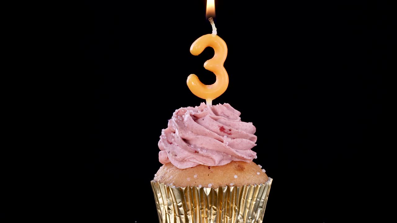 A single cupcake with pink frosting and a lit number three candle burns steadily against a black background, illuminated by soft, focused lighting