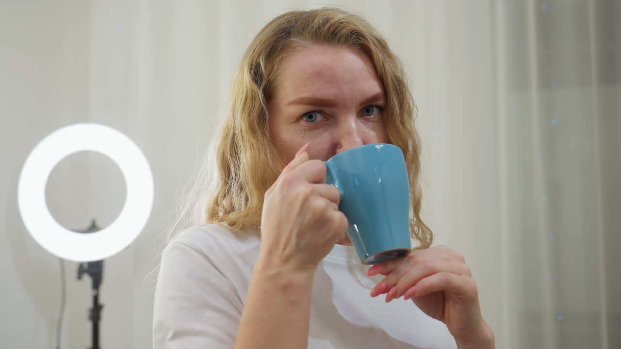Blond woman with nose ring sipping hot tea from blue cup while standing in softly lit room with vanity mirror and ring light, expressing peaceful moment of calm and enjoyment during indoor routine