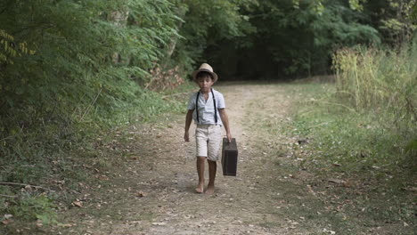 boy walking barefooot towards the camera wearing vintage clothes a hat and an old suitcase in the forest