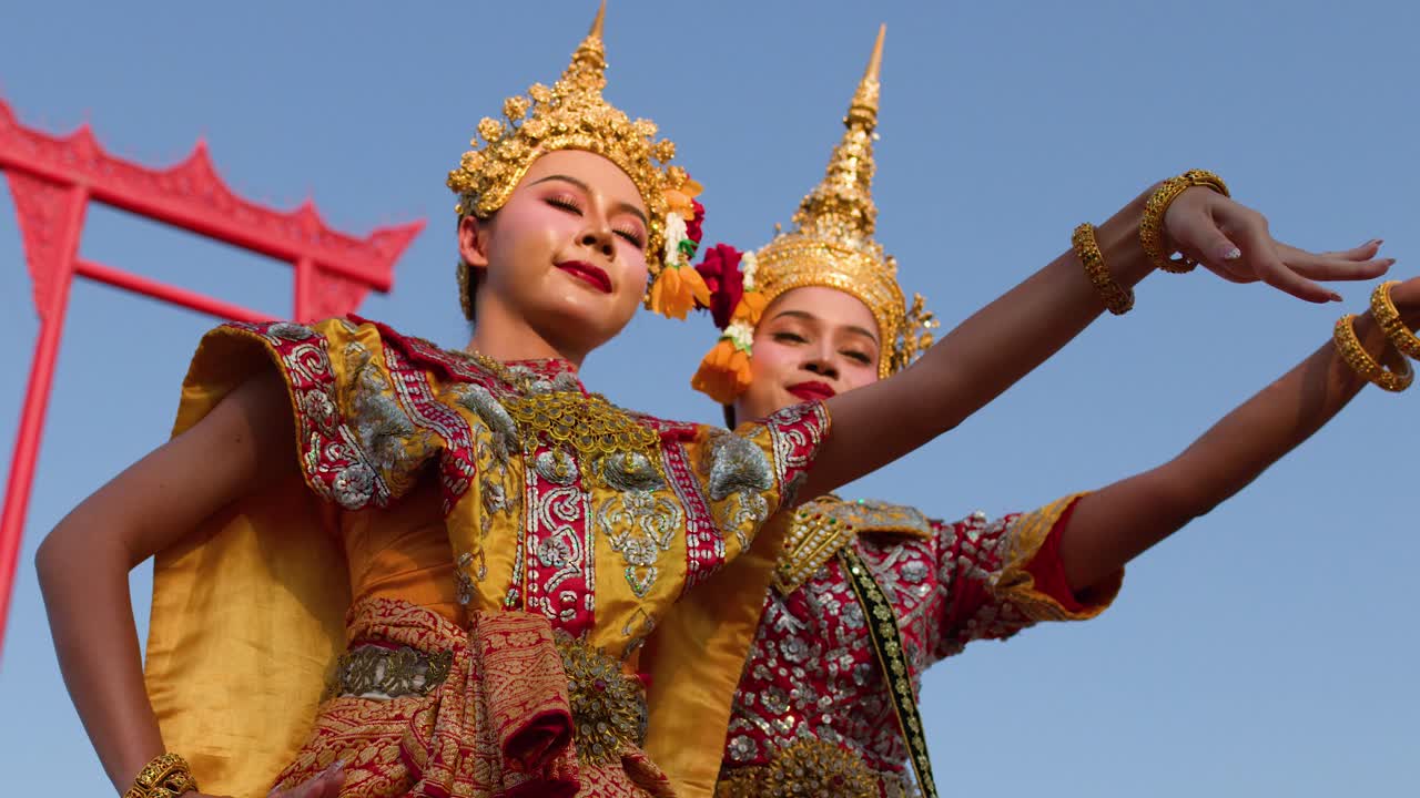 Dua penari wanita dengan kostum emas berhias menampilkan gerakan tangan yang anggun di luar ruangan di bawah sinar matahari terang