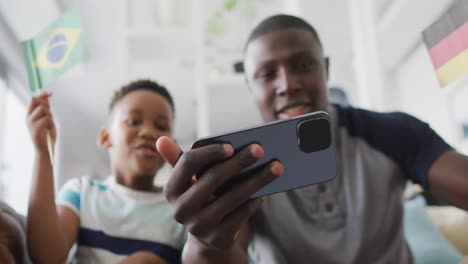 Video-of-happy-african-american-father-and-son-sitting-on-sofa-and-watching-match-on-smartphone