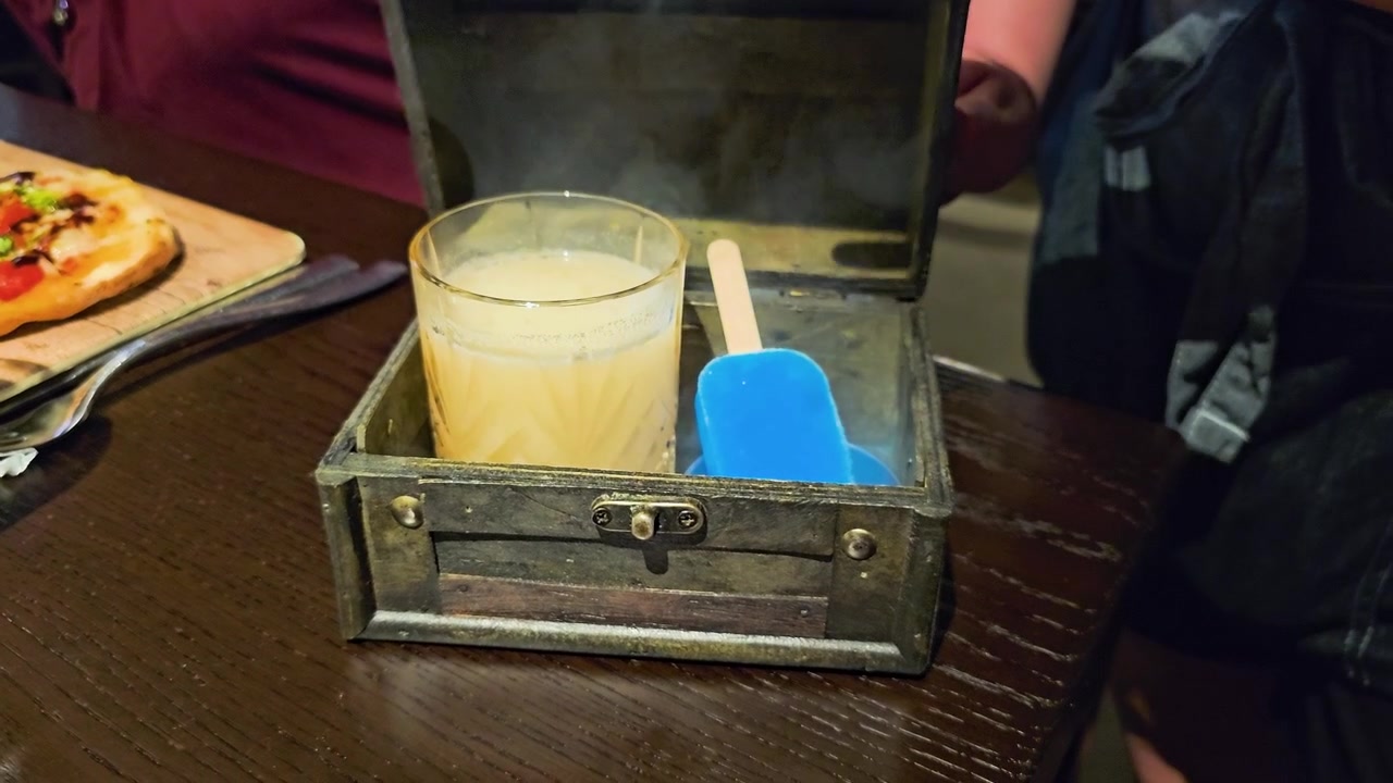 Una mano chiude una rustica scatola di presentazione in legno sopra un cocktail fumante e una guarnizione di ghiacciolo blu su un tavolo di ristorante, con legno scuro, illuminazione soffusa da bar e un'atmosfera da pranzo drammatica.