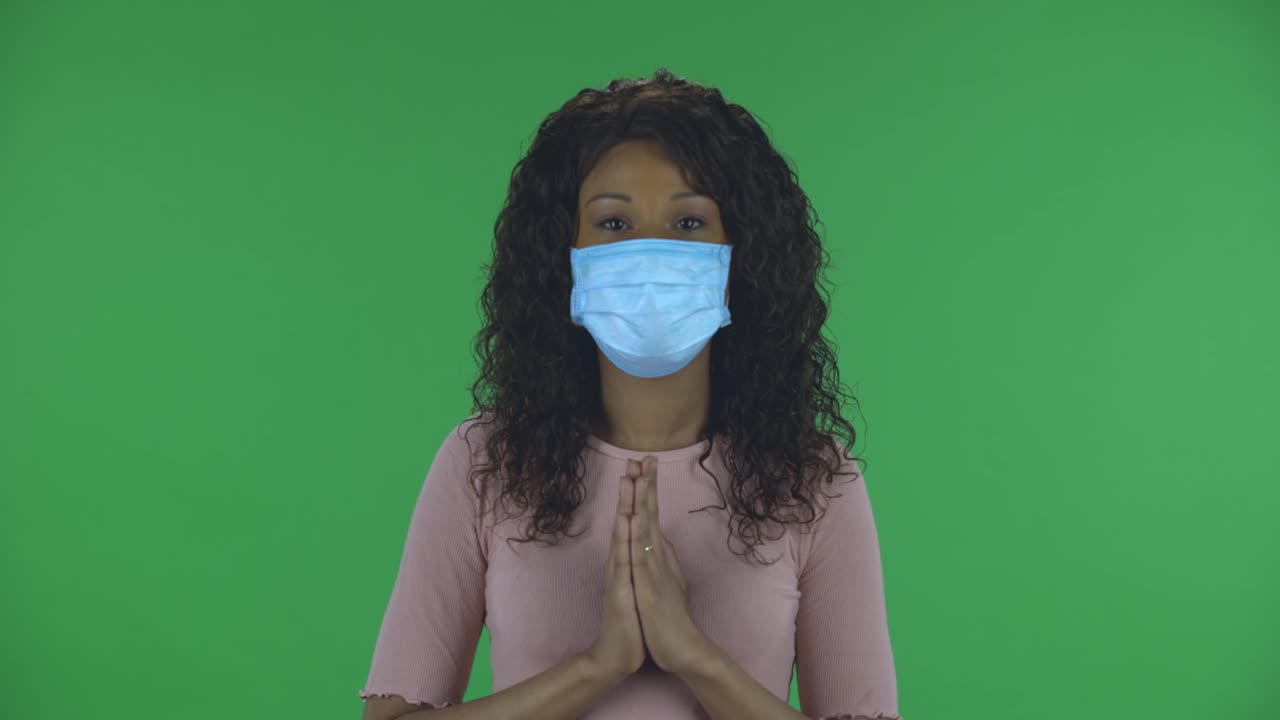 retrato de una hermosa mujer joven afroamericana con máscara médica está mirando directamente y reza con las manos juntas palma a palma. brunette ardiente con cabello ondulado en vaqueros y una blusa beige en una pantalla verde en el estudio. protección de la salud coronavirus