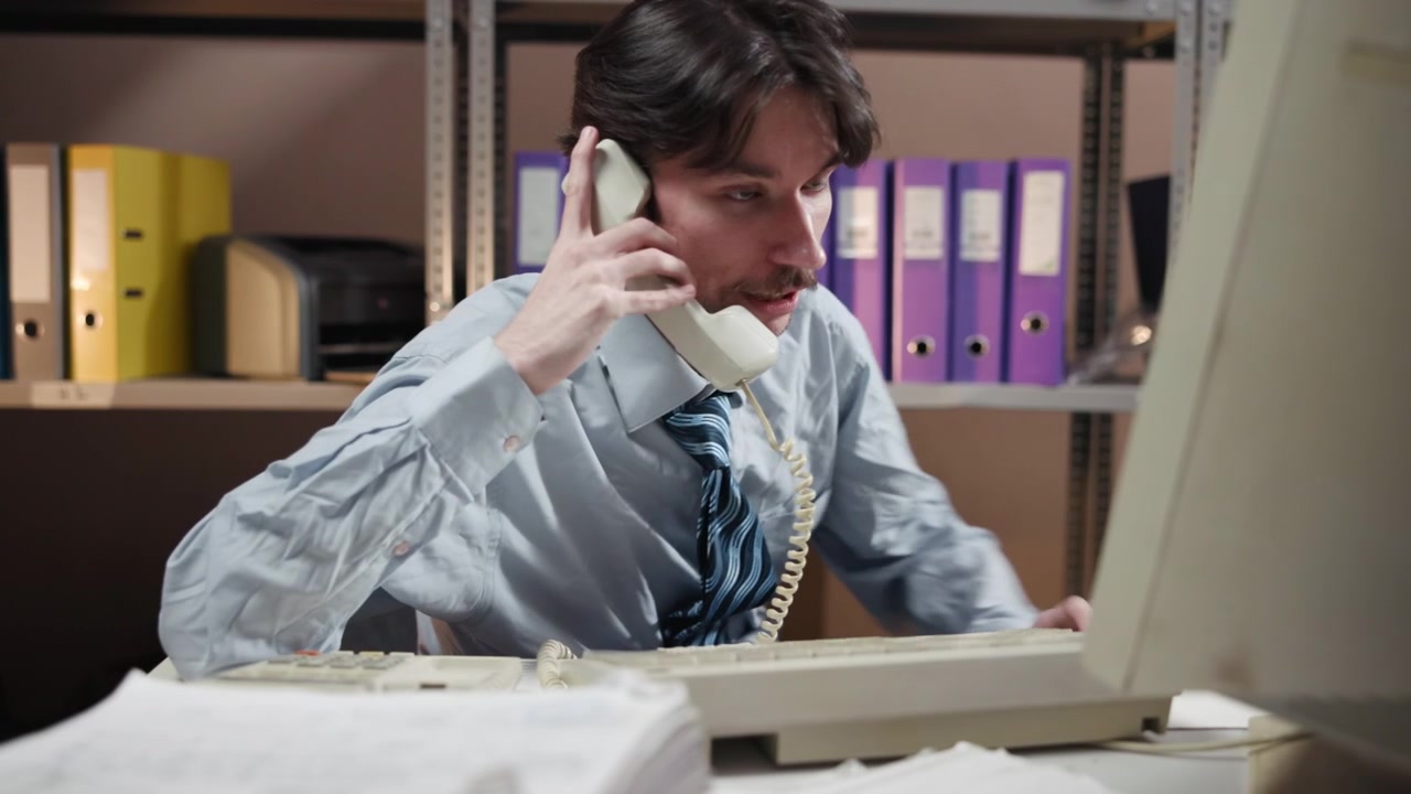 Un impiegato stanco si appoggia all'indietro a una scrivania vicino a un monitor di computer, con scaffali pieni di raccoglitori e faldoni sullo sfondo in un interno d'ufficio poco illuminato e ordinato.