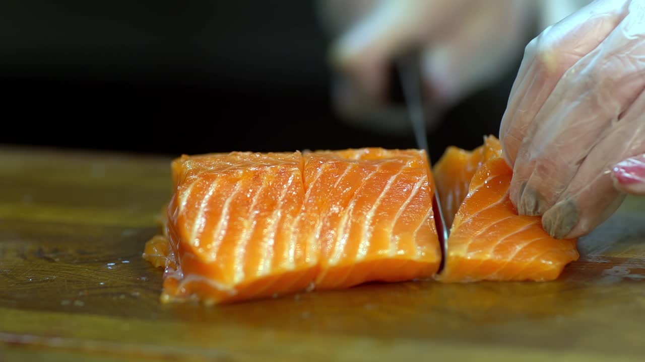 A salmon fillet is cut into pieces by hands wearing hygienic plastic gloves