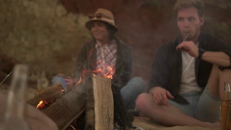 Group-of-young-friends-sitting-by-the-fire-on-the-beach,-drinking-beer-and-grilling-marshmallows.-Slow-Motion-shot