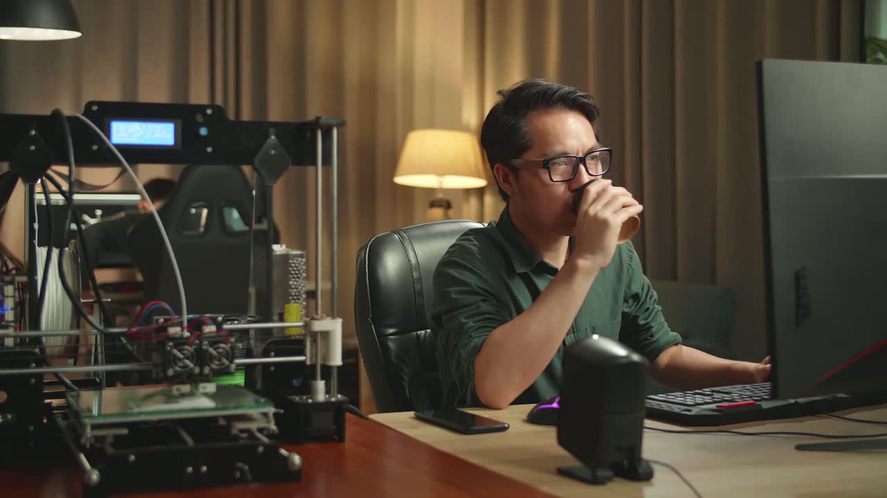 Asian Man Drinking Coffee While Works On Personal Computer And 3D Printer In Home Office, He Works At Night