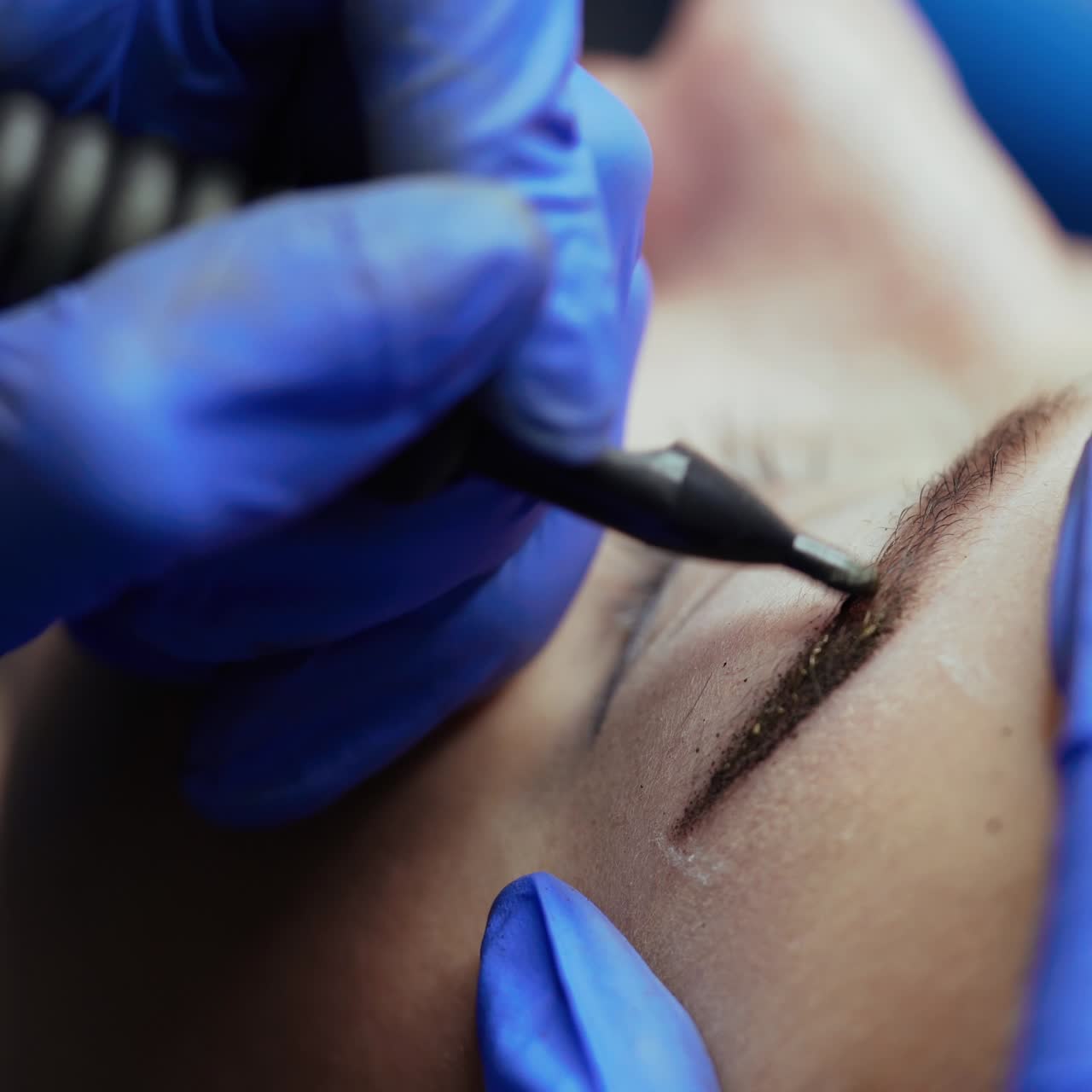 Beauty master makes eyebrow tattoo in blue gloves with the use of a special needle and a pistol in the salon. Close-up
