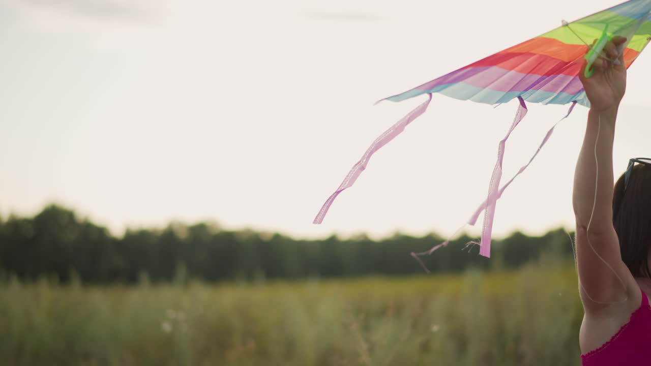 Mujer negra caminando por una carretera rural con un vestido rosa junto a una cometa arcoíris caída, sonriendo, preparándose para lanzarla, cielo dorado, borde cubierto de hierba, ambiente relajado y seguro, poca profundidad de campo cinematográfica