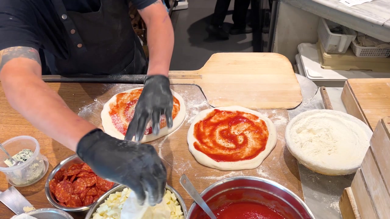 Chef con guanti neri sparge farina e aggiunge condimenti all'impasto della pizza su un bancone di legno
