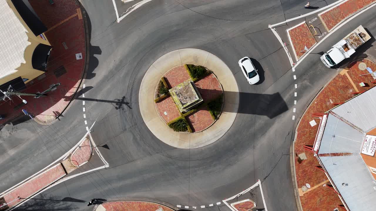 Overhead drone footage captures cars, trucks, and buses moving through a sunlit roundabout in Coonabarabran, New South Wales, with clear shadows and urban surroundings