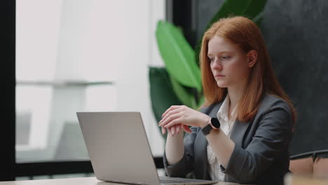 A-young-red-haired-business-woman-looks-thoughtfully-at-the-screen-and-brainstorms.-Watch-and-think-about-problems-looking-out-the-window.-Thoughtful-business-woman