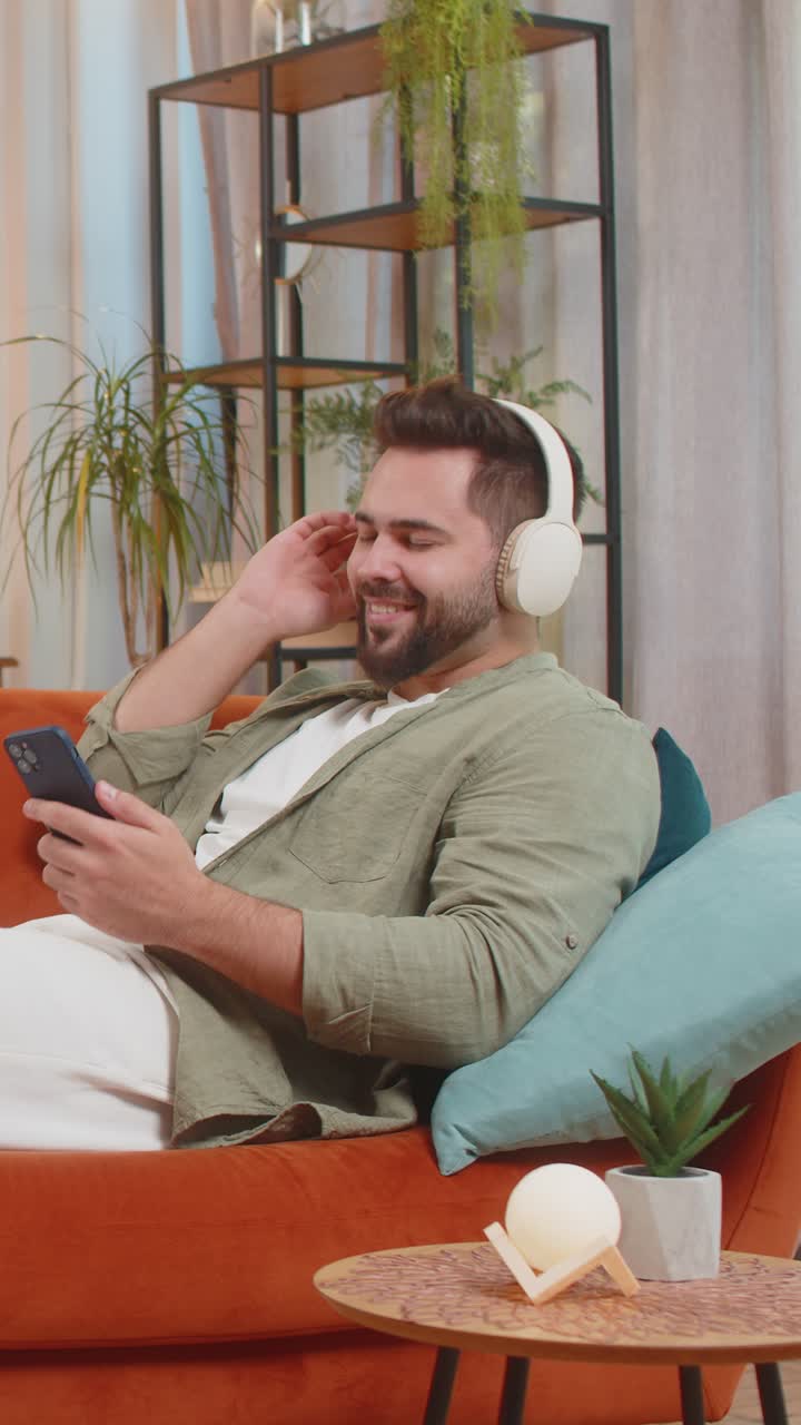 joven feliz y encantado con auriculares inalámbricos usando un teléfono inteligente relajándose sentado en el sofá en casa