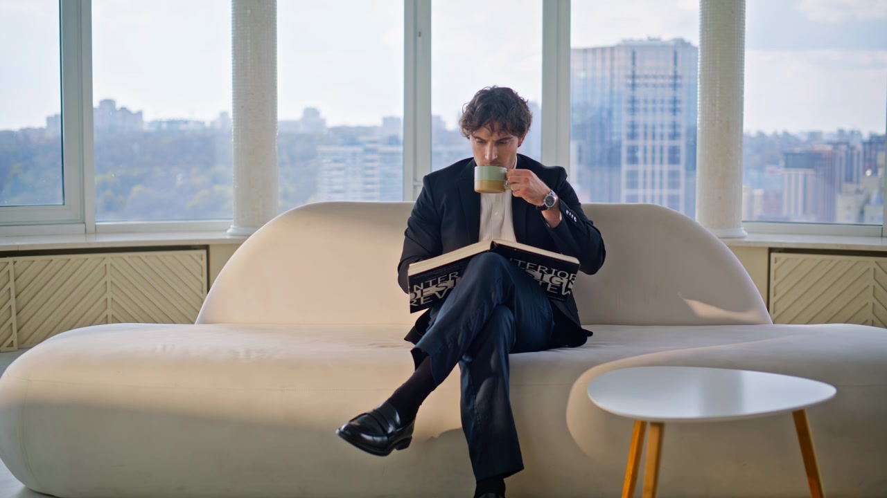 Elegant mann leser designbok og drikker kaffe på sofaen i en luksusleilighet. Nærbilde.