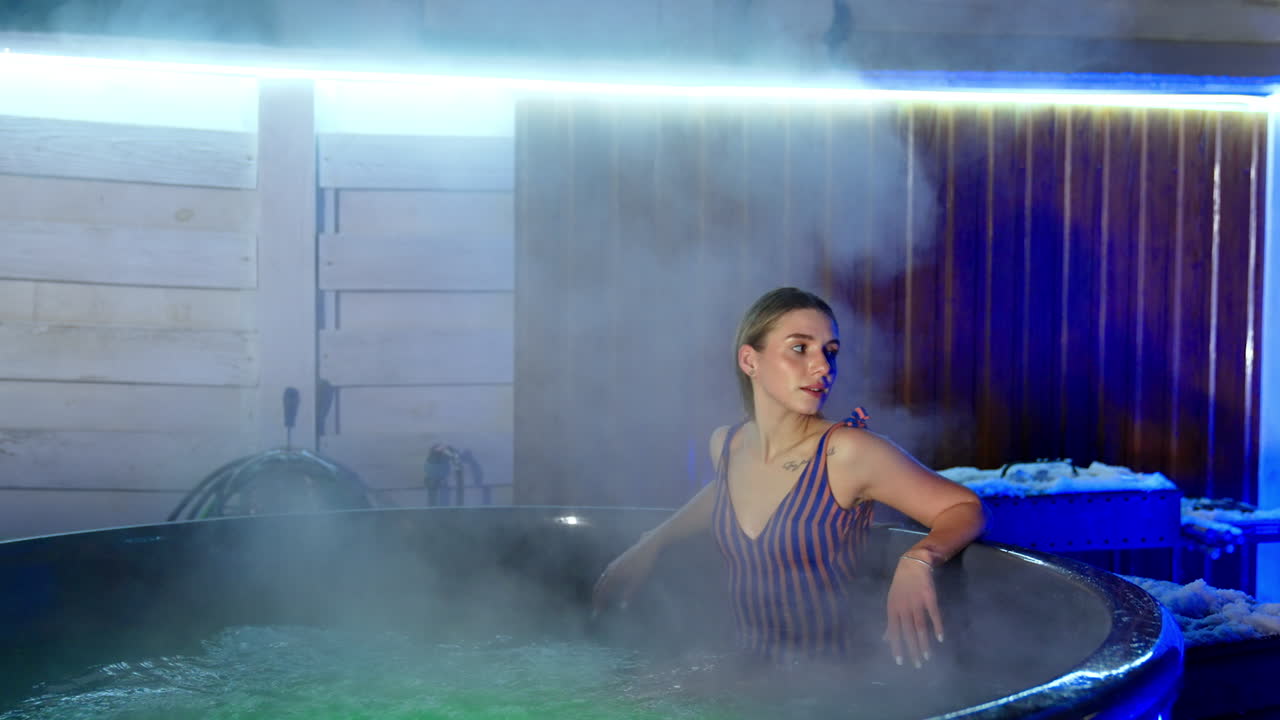 Good-looking Caucasian woman in stripy swimsuit sits on the edge of the Jacuzzi. Lady splashes water with her hand and then deeps in hot tub.