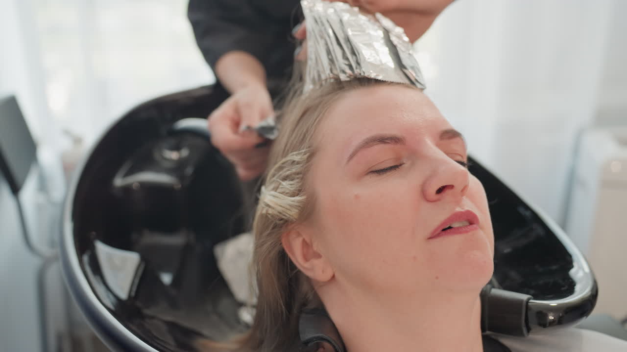 Donna caucasica al lavatesta di un salone di bellezza mentre riceve la colorazione e il risciacquo dei capelli con la stagnola. La stilista applica i colpi di sole con la stagnola di alluminio ed effettua il risciacquo in un lavandino di porcellana nero mentre la cliente indossa un mantello protettivo e si rilassa.
