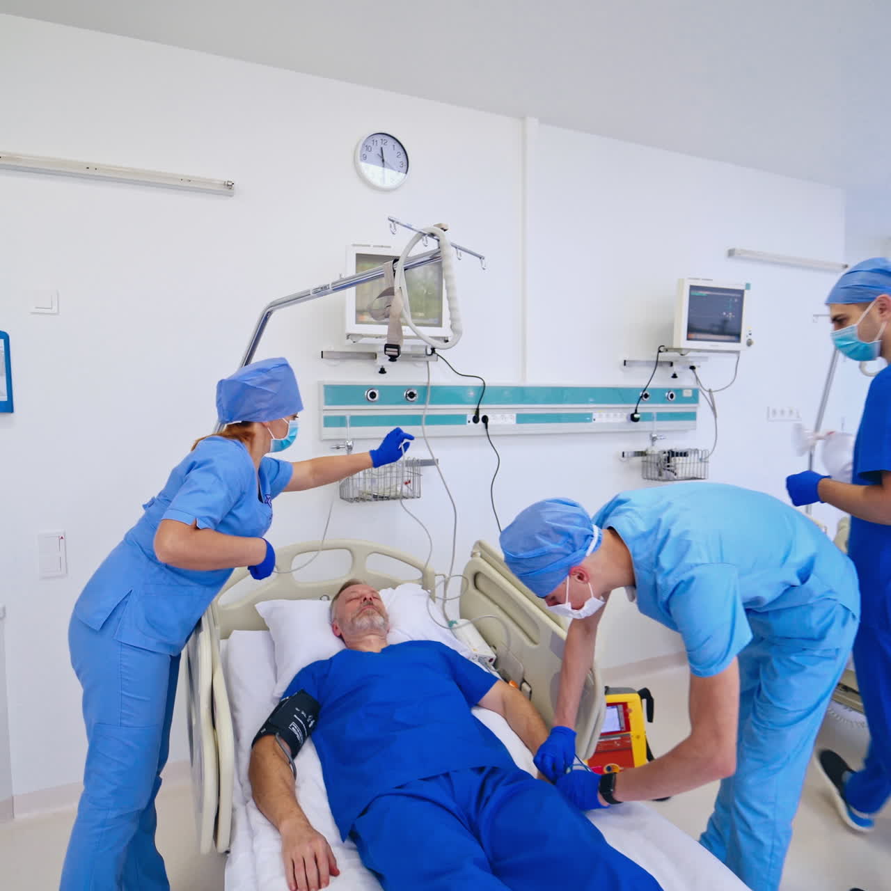 Male patient is lying on the bed in emergency room. Medical workers in masks and protective clothing prepare equipment for saving patient's life in modern clinic.