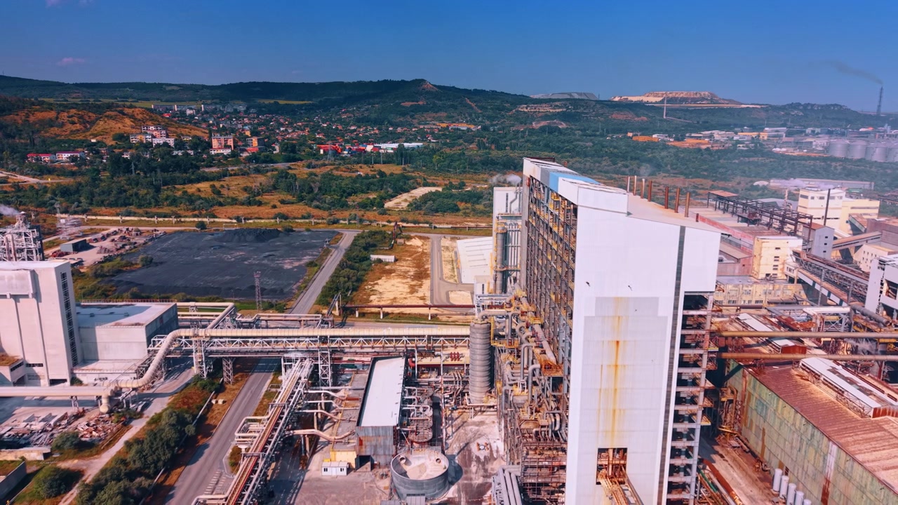 Modern industrial building with external pipeline network near town hills. High angle perspective of factory white facade, complex tube lines, green landscape and distant village.
