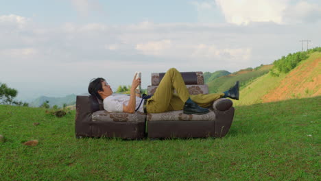 asiatic male student laid on vintage sofa couch in outdoor scenic location with green natural mountain valley landscape in a sun day of summer reading a book