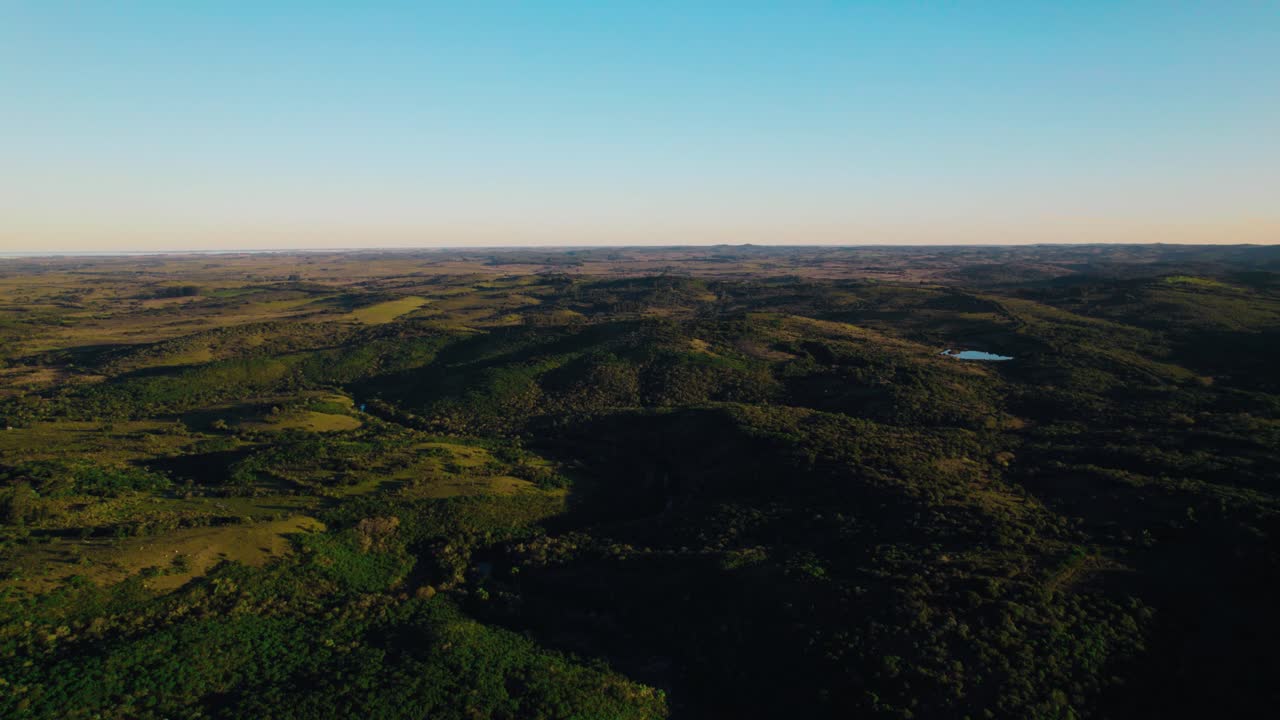 An aerial drone view capturing the vast, tranquil landscape of the Uruguayan countryside at sunrise. Features rolling hills, green fields, forests, and a clear blue sky, suggesting peace and nature