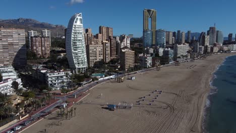 Vuelo-Aéreo-Sobre-La-Playa-De-Arena-En-Benidorm-Con-Edificios-Modernos-Llamados-Delfin-E-Intempo-Tower-En-Segundo-Plano