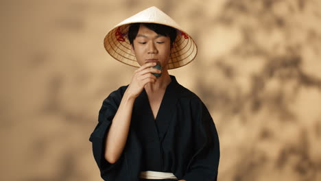 young asian man in black traditional costume and conus hat drinking tea and smiling at camera