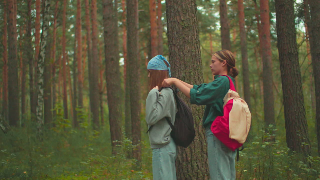 In a serene forest, a young woman in a red backpack helps her friend wearing a blue scarf adjust her black bag, they share a warm moment, smiling at each other, before continuing their hike