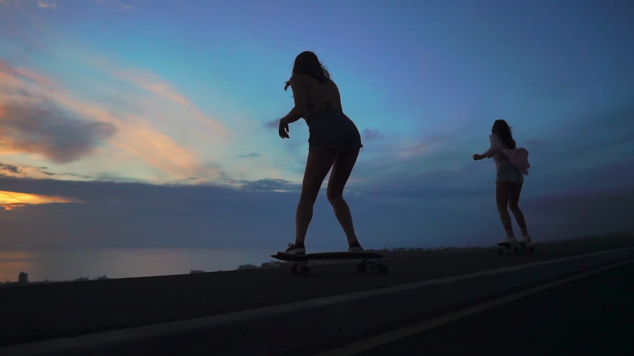 In slow motion, two friends skate along a road at sunset, with mountains and a stunning sky as the backdrop. They're wearing shorts