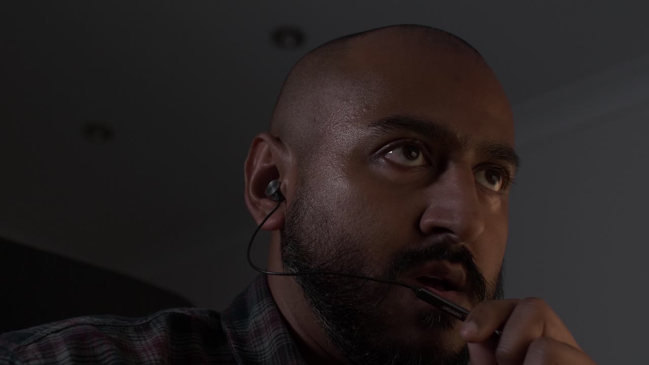 UK Asian Male Talking On Earphone Microphone Indoors In Dimly Lit Room