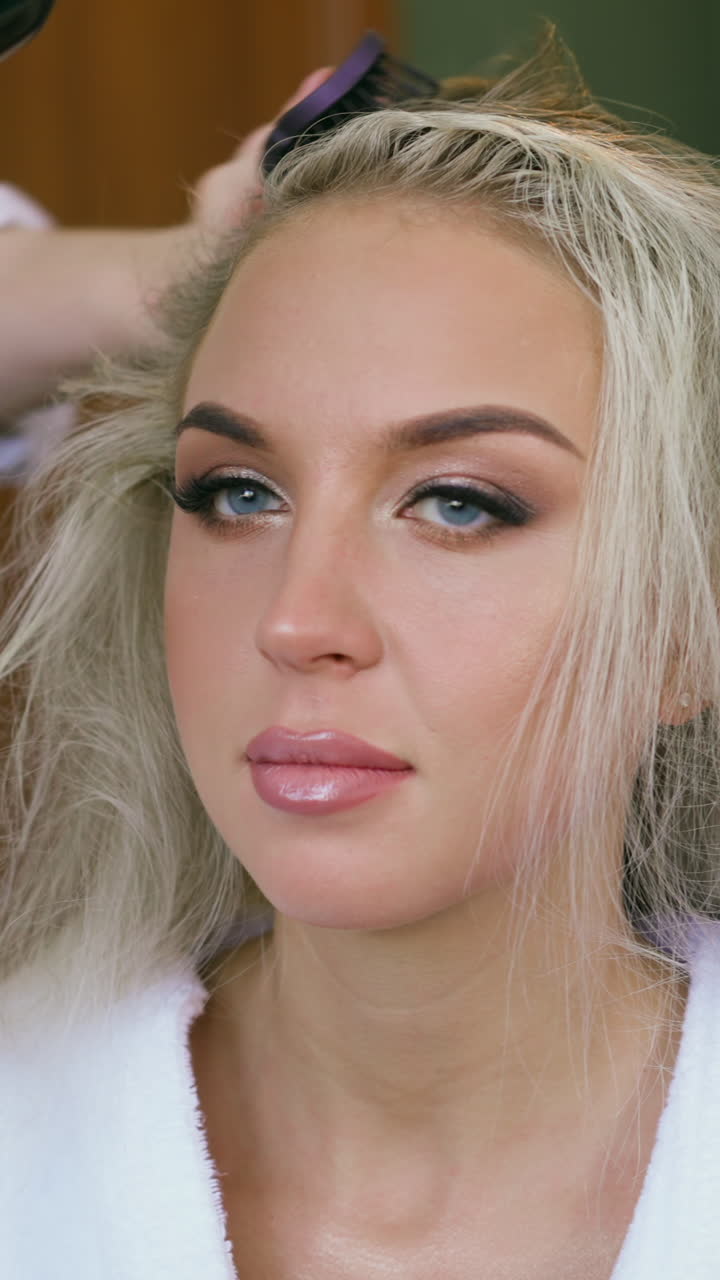 Pretty young blonde woman with festive makeup sits on chair while skilled stylist does hairstyle with brush and blow dryer in beauty salon