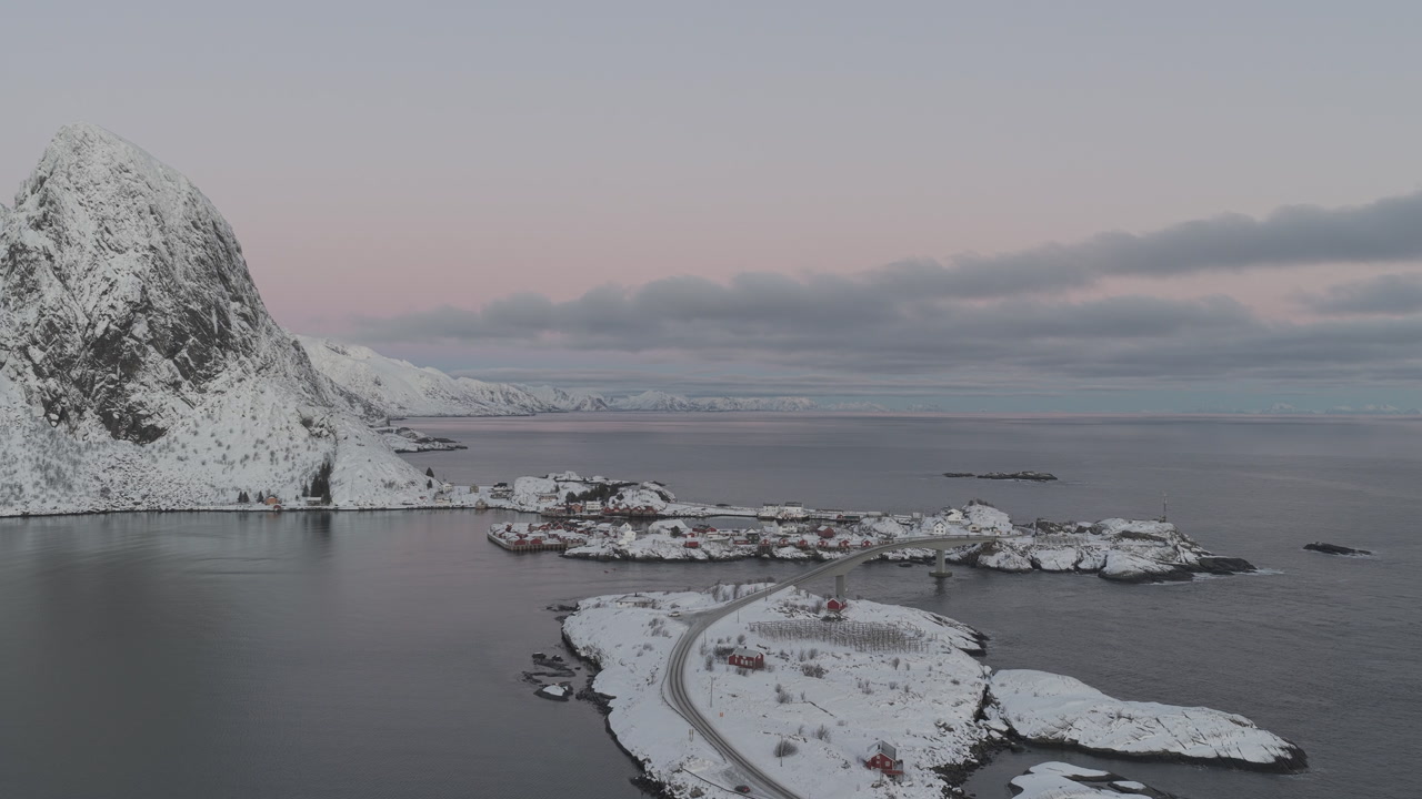 A stunning cinematic aerial shot of the snowcapped mountains and coastal islands of Lofoten, Norway, featuring a winding road and bridge during a cold winter dawn.