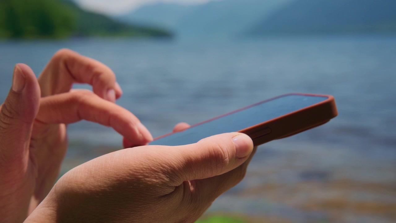 Mani che usano il telefono vicino a un lago. Lo sfondo mostra acqua calma e colline verdi distanti. La persona appare rilassata e concentrata sul dispositivo. Ideale per temi di viaggio, tecnologia o comunicazione all'aperto.
