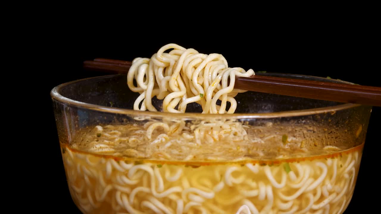 Chopsticks rotate above a clear glass bowl, lifting a bundle of instant noodles in broth. Studio lighting, black background, smooth camera tracking