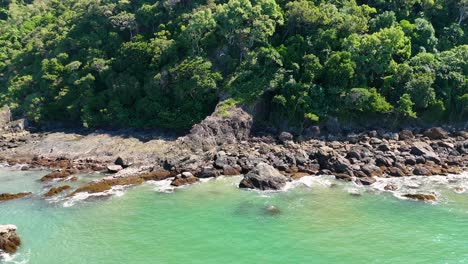 drone glides above a deserted rocky shoreline bordered by lush rainforest and turquoise water under bright daylight, capturing sweeping coastal scenery