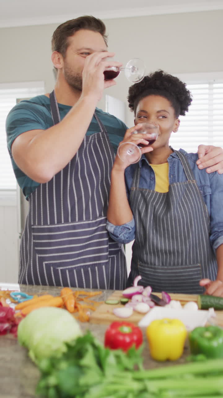 video vertical de una pareja feliz y diversa preparando comida en la cocina y bebiendo vino