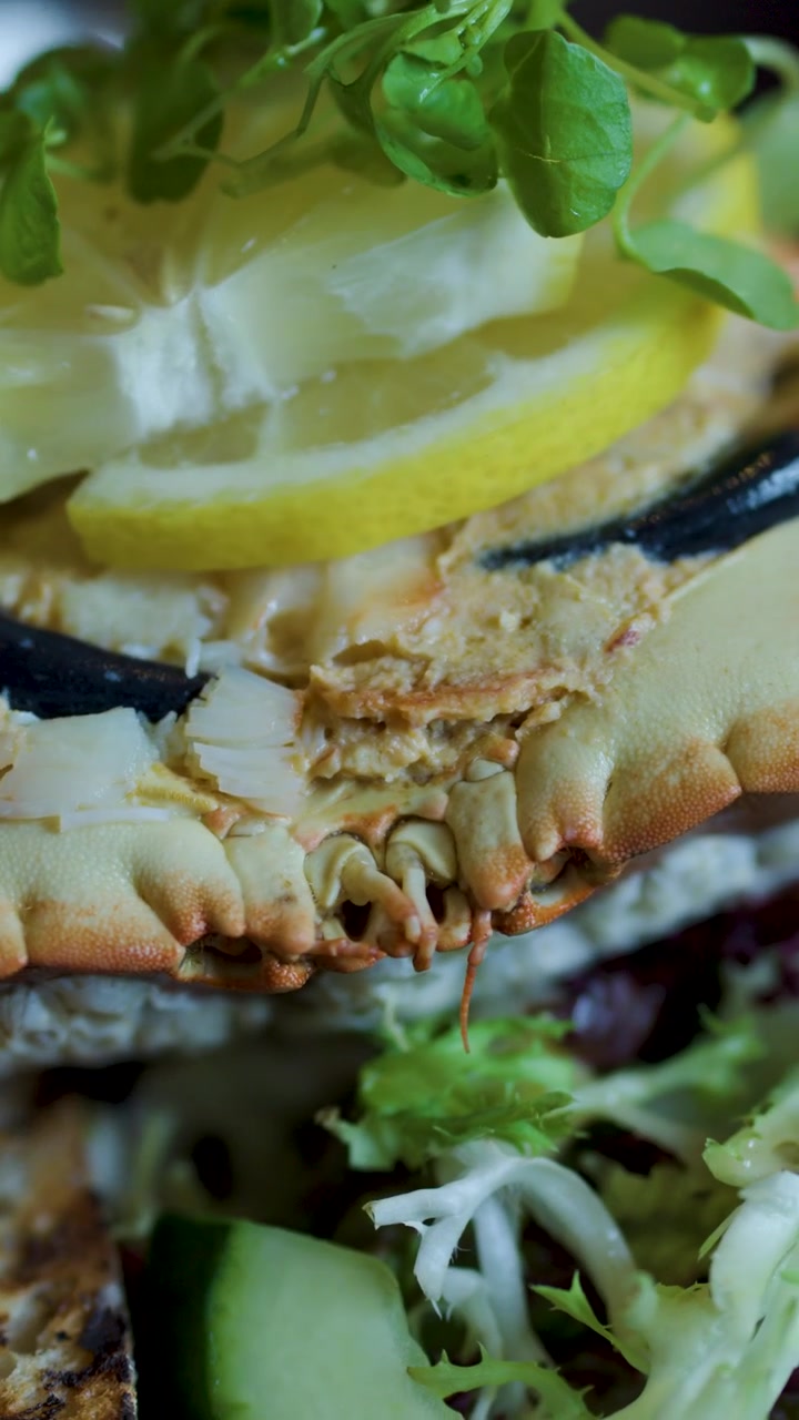 Kamera bergerak perlahan melintasi kue kepiting yang disajikan di piring dengan lemon, sayuran hijau segar, dan hiasan mentimun.