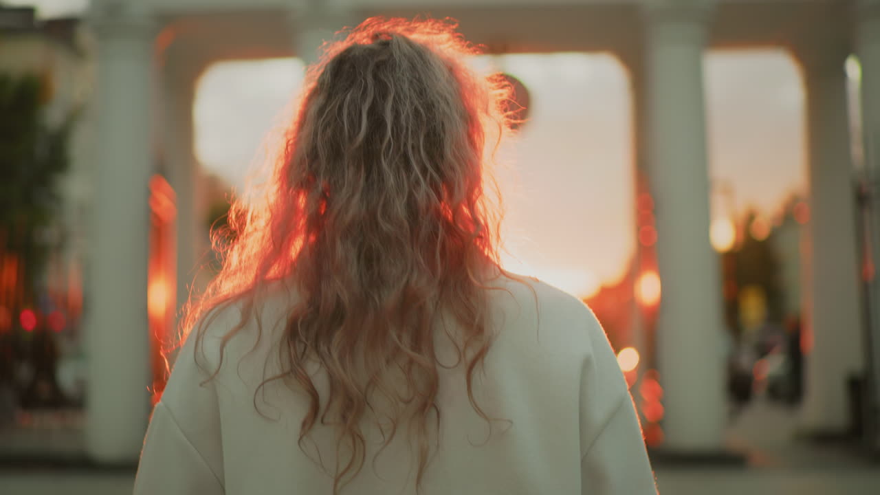 back view of woman with curly hair walking toward sunlit archway in urban setting as glowing red sunlight illuminates her hair with blurred motion of cars and people in background
