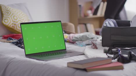 portátil de pantalla verde con una maleta de embalaje de persona en la cama en casa para las vacaciones de verano