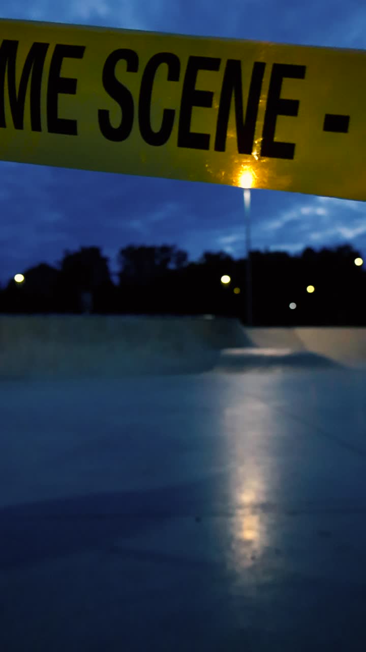 Gorgeous cinematic close up or closeup footage of a yellow vibrant colored Crime Scene Do Not Enter tape hanging in front of a dark evening skatepark with street lamps back lighting the ribbon tape