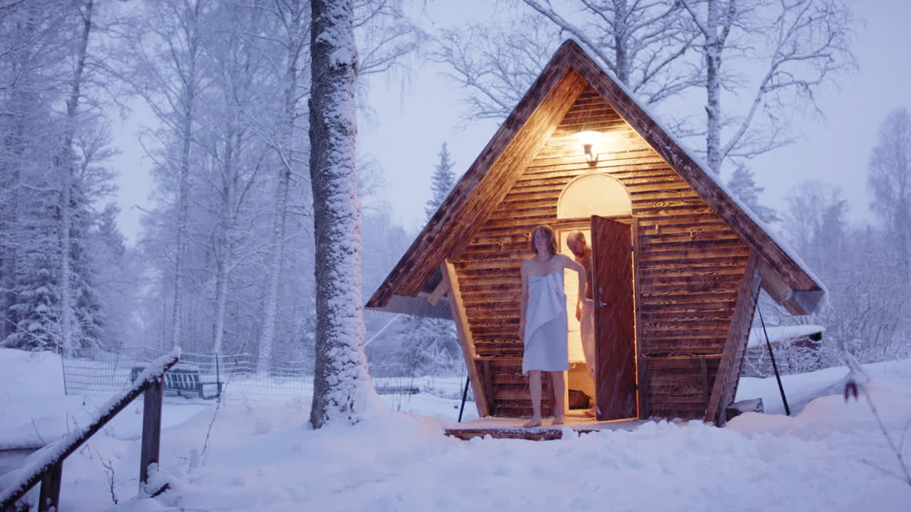 Couple exits wooden sauna to sit outside surrounded by snowy winter wonderland