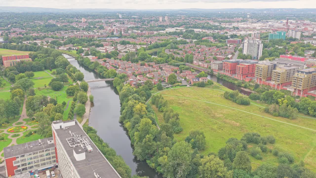 Immagine aerea statica mostra i prati di Salford e il fiume Irwell, con la sagoma curva del Trinity Bridge di Calatrava che collega Salford a Manchester tra un parco verde e uno sfondo urbano.