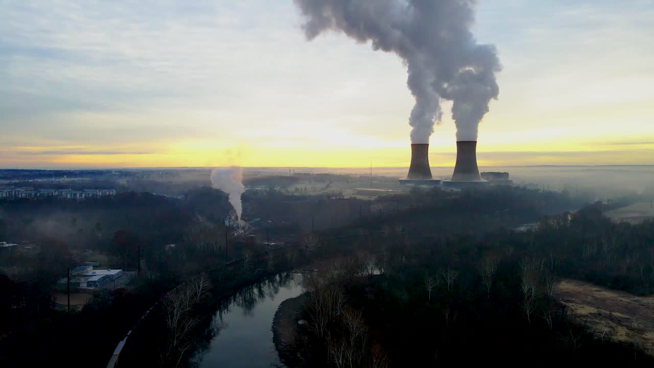 Un impianto nucleare all'alba, con le torri di raffreddamento che emettono vapore nel cielo dell'alba. C'è un treno merci in basso a sinistra nell'inquadratura