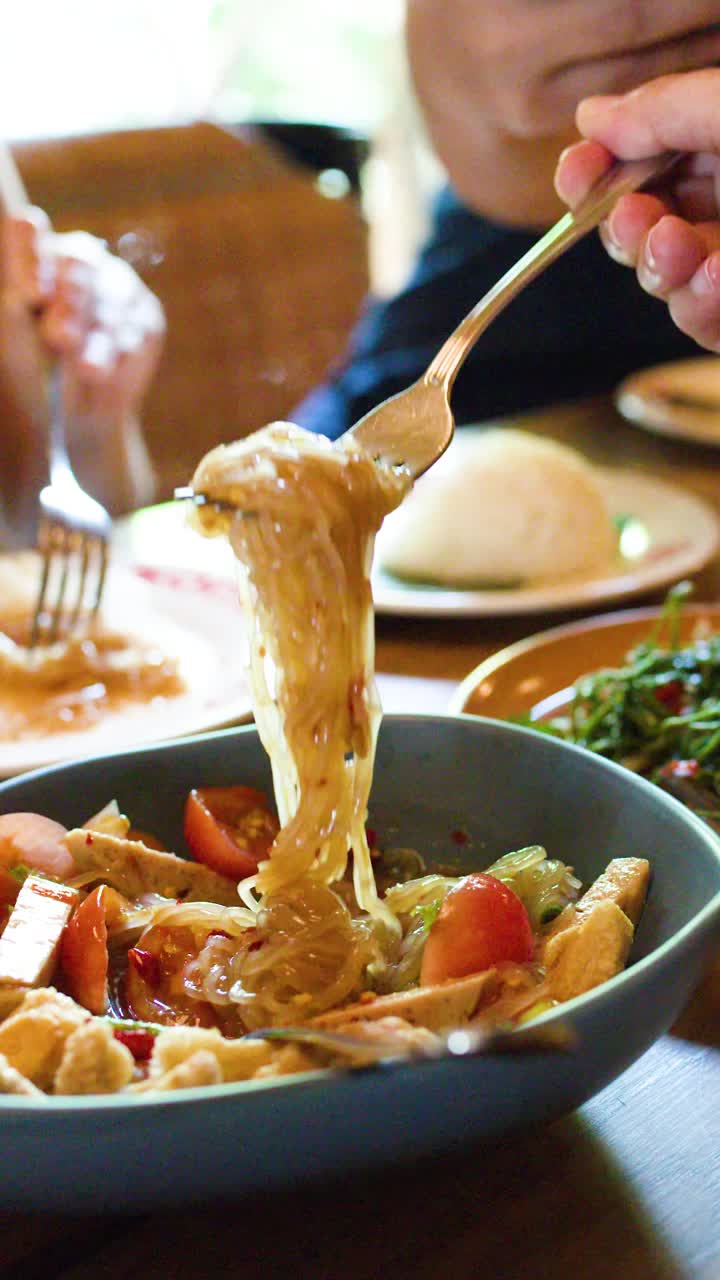 Primo piano di una mano che mescola un'insalata piccante di vermicelli di riso tailandesi, illuminata da luce naturale, in un ambiente da pranzo informale.