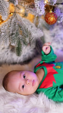 sweet little boy in green elf costume lies on white plaid under beautifully decorated christmas tree. cute child tosses his feet actively and looks into camera. vertical video