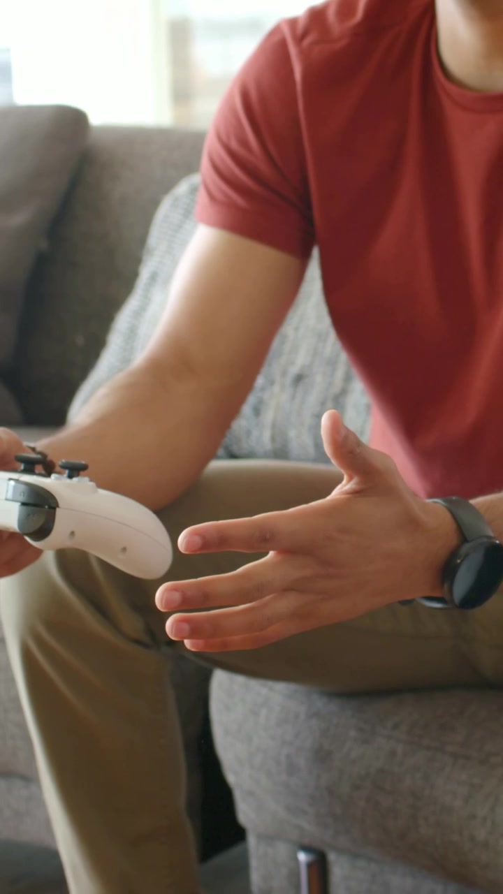 Video verticale: Uomo in maglietta rossa che si sistema sul divano, sintonizza un gamepad bianco e solleva un visore VR per giocare.